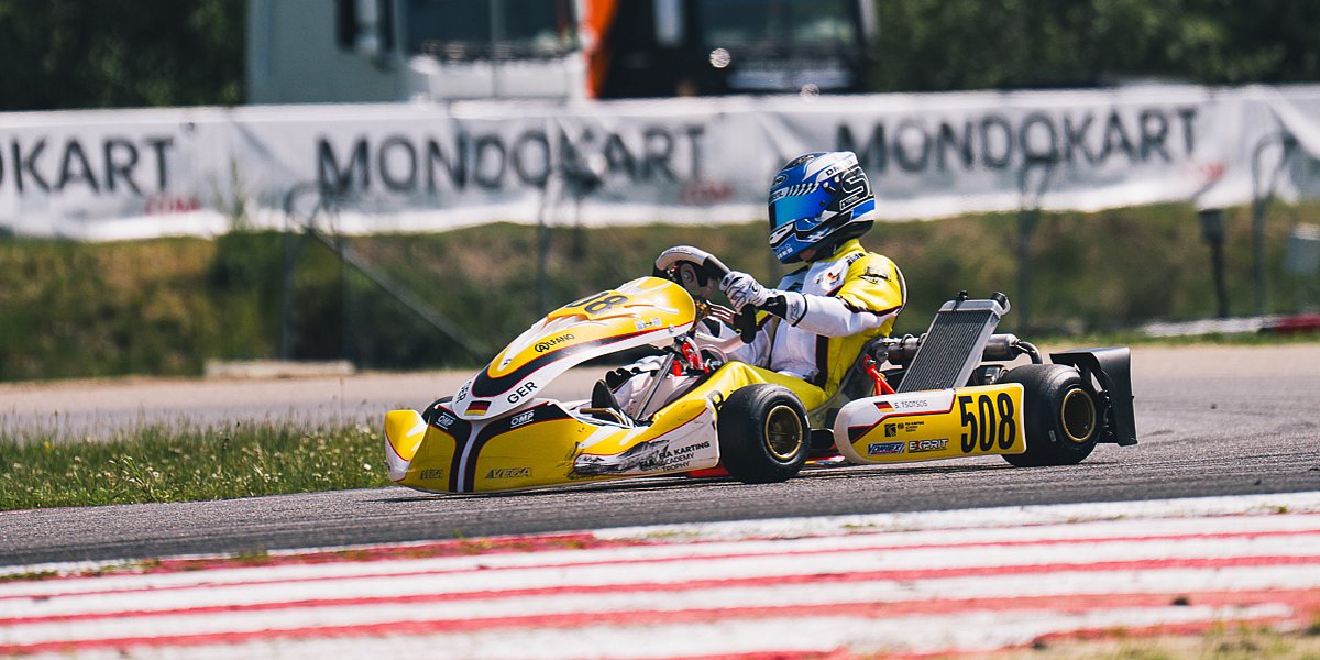 Stavros Tsotsos Francia fährt im gelben Kart durch eine Kurve der FIA Karting Academy Trophy, im Hintergrund sind MONDOKART-Banner und Transportfahrzeuge zu sehen.