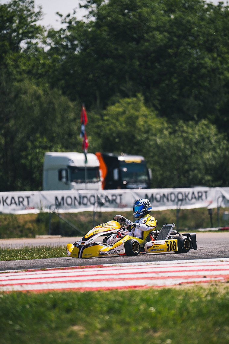 Stavros Tsotsos Francia fährt im gelben Kart durch eine Kurve der FIA Karting Academy Trophy, im Hintergrund sind MONDOKART-Banner und Transportfahrzeuge zu sehen.