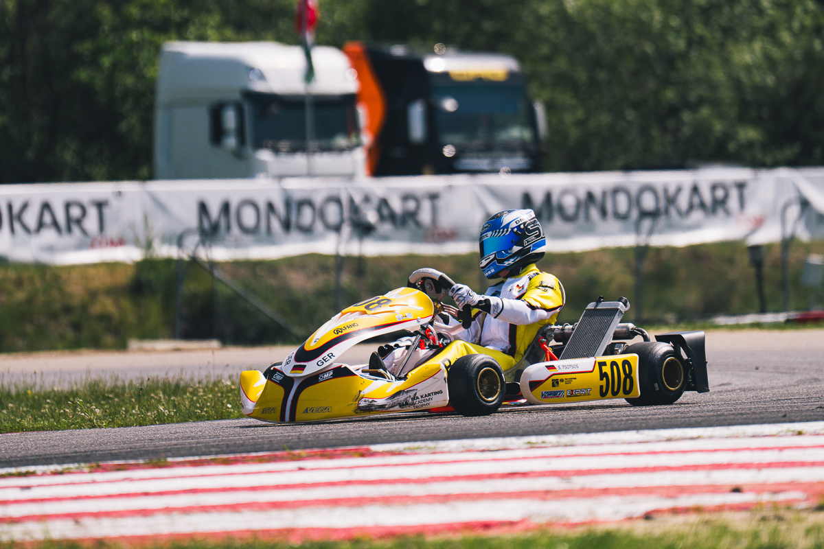 Stavros Tsotsos Francia fährt im gelben Kart durch eine Kurve der FIA Karting Academy Trophy, im Hintergrund sind MONDOKART-Banner und Transportfahrzeuge zu sehen.