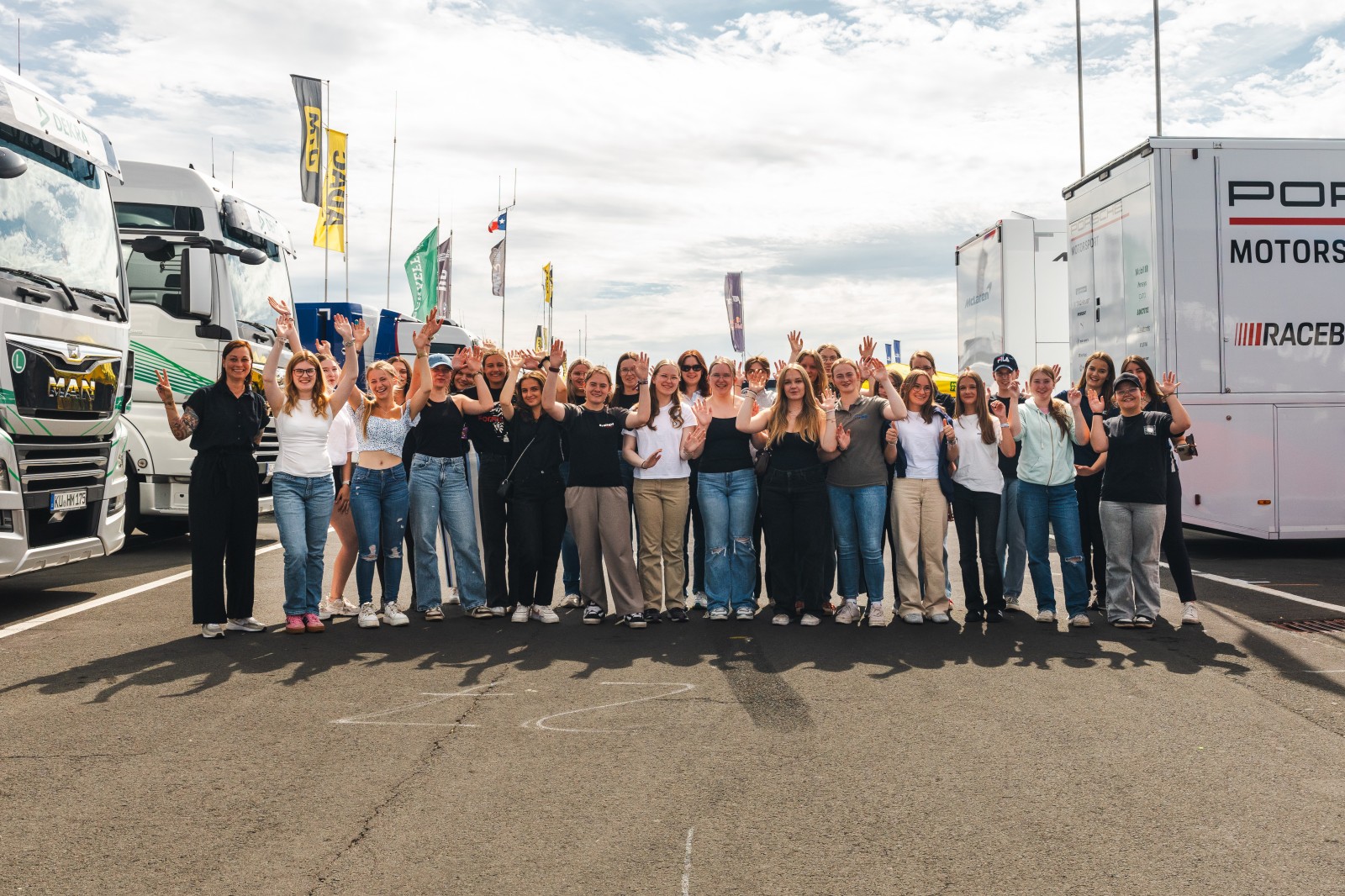 Discover Motorsport – Young Women’s Edition 2025 Gruppenfoto von Teilnehmerinnen der Discover Motorsport – Young Women’s Edition 2025 am Nürburgring mit erhobenen Händen vor den DTM-Teamtruckern, dazu das Event-Logo und der Hinweis „Am 23. Juli in Oschersleben – jetzt anmelden“.