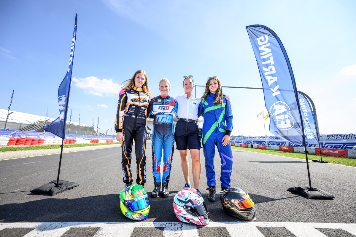 3 Fahrerinnen in Rennanzügen und eine Frau der FIA auf der Rennstrecke, vor ihnen liegen ihre Helme, an der Seite Beach Flags zu FIA Karting